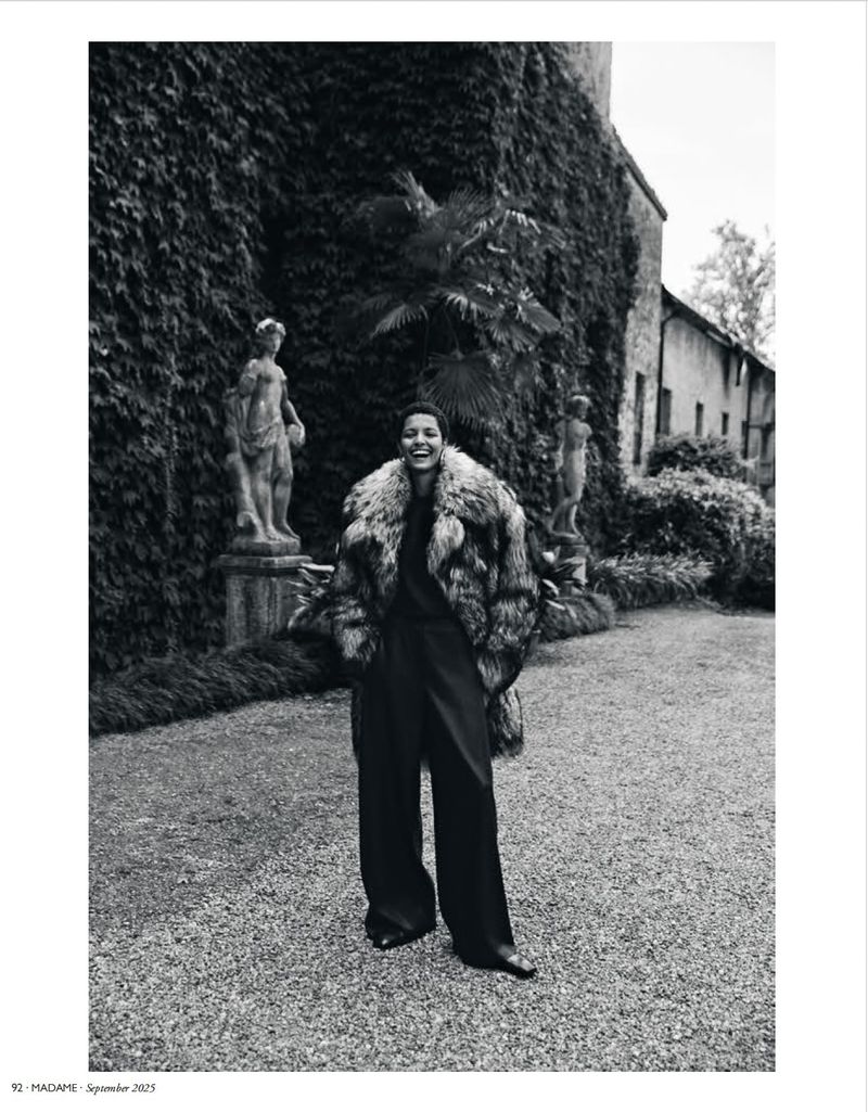 Woman poses in fur coat with statues and a building covered in ivy. Black and white fashion editorial in a garden setting.