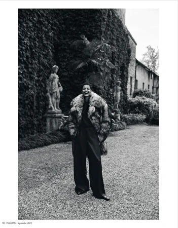Woman poses in fur coat with statues and a building covered in ivy. Black and white fashion editorial in a garden setting.