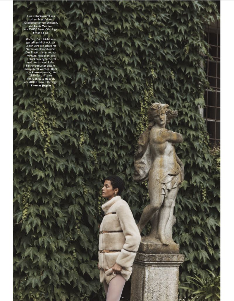 A woman in a fur jacket stands by a stone sculpture in a garden with lush greenery, captured in a fashion editorial setting.