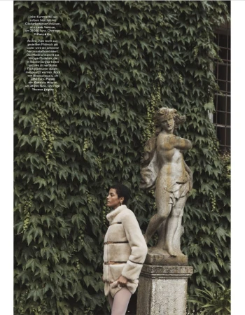 A woman in a fur jacket stands by a stone sculpture in a garden with lush greenery, captured in a fashion editorial setting.