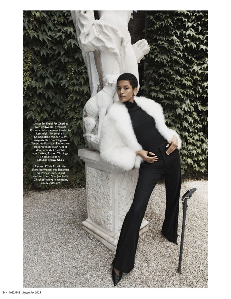 A woman in a black turtleneck and black pants stands next to a stone statue, wearing a white fur coat, in a garden setting.
