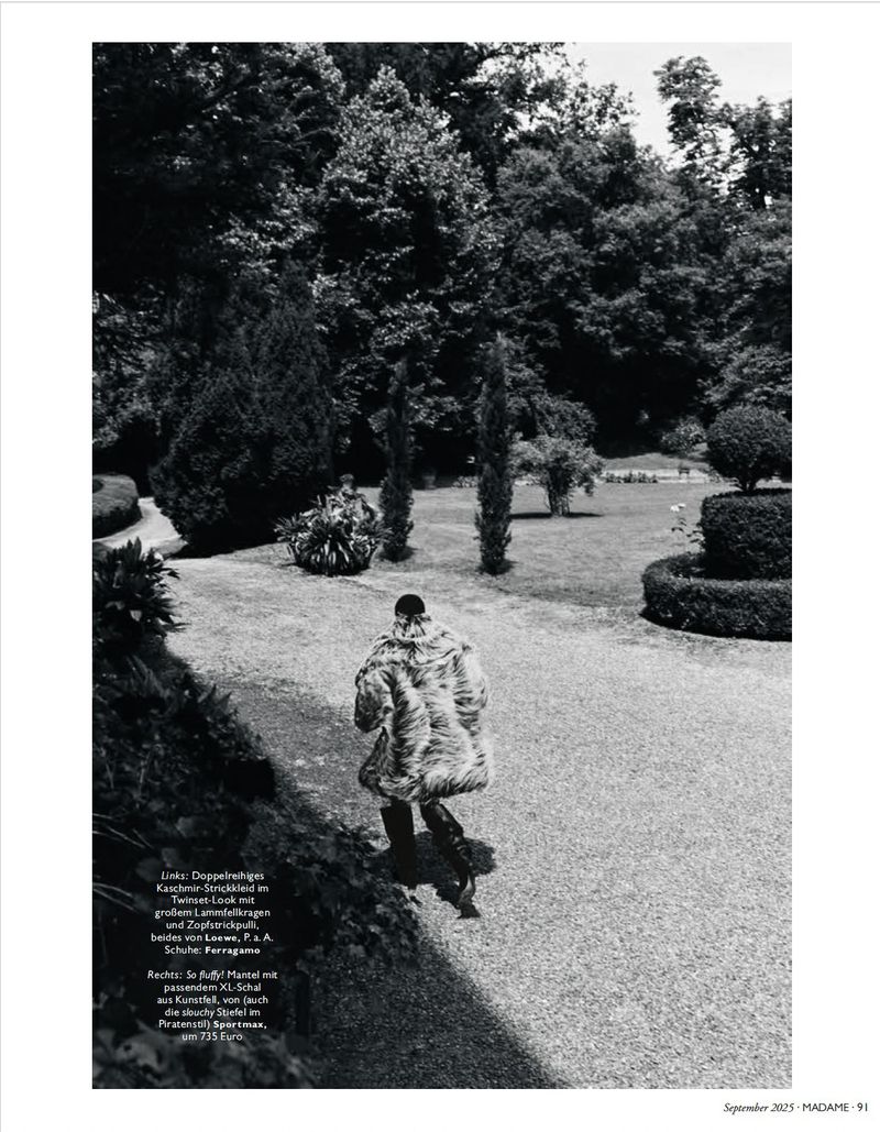 Person walks on a stone path through lush green park, wearing fur coat and dark trousers with trees and foliage in background.