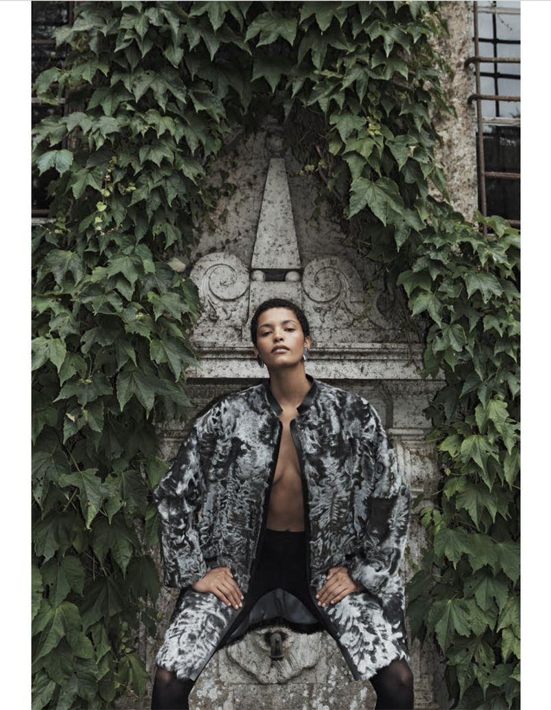 Fashionable adult female model poses in a patterned coat outdoors against a stone fountain with green ivy, eyes looking to the front.