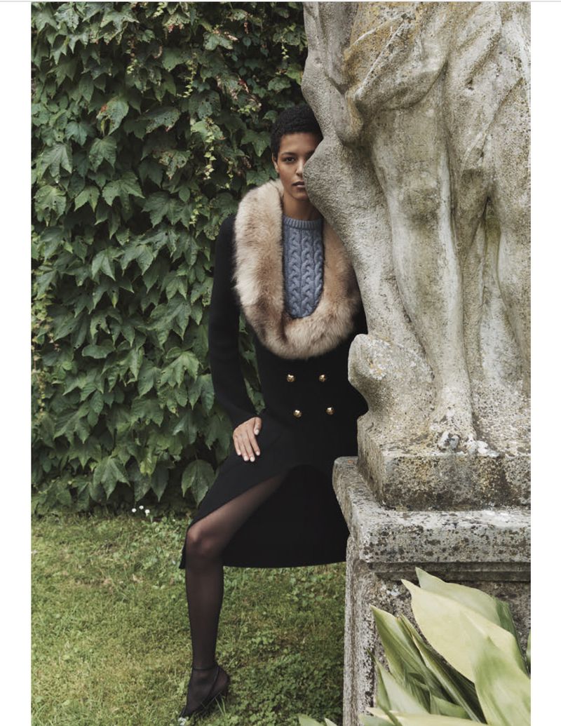 Fashionable woman in black coat with fur collar poses in garden by stone structure, capturing a blend of nature and style.