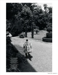 Model walking away on a gravel garden path wearing a textured fur coat and dark boots in a black and white fashion editorial image.