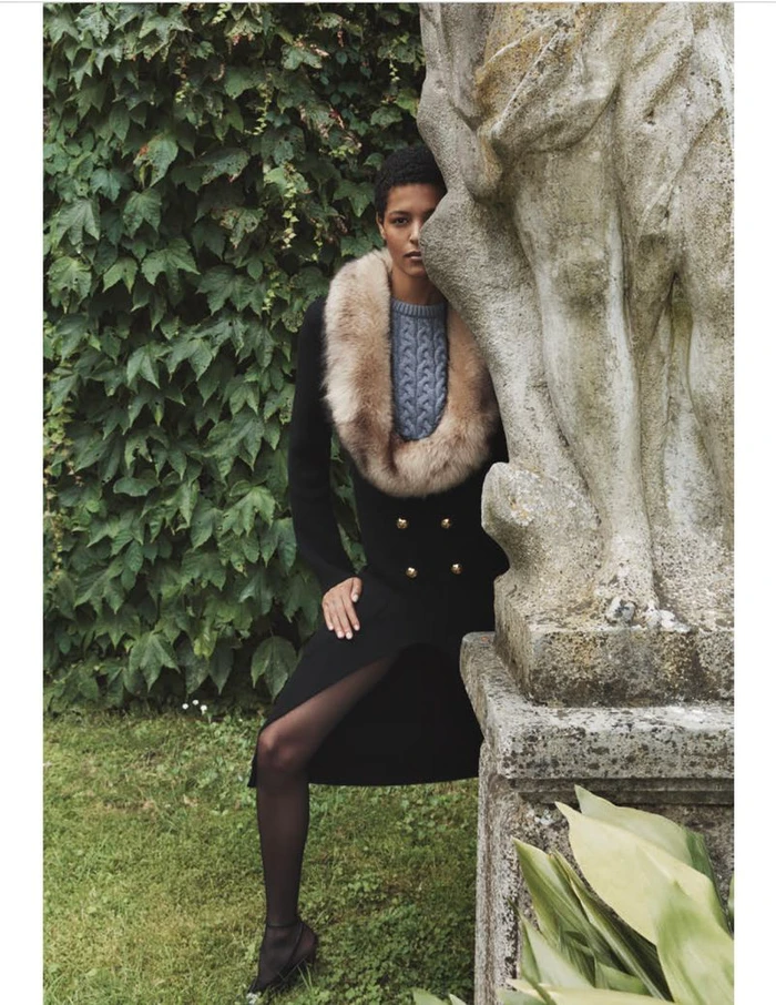Model Laiza de Moura wearing a black coat with a fur collar standing beside a stone statue in front of a leafy hedge.