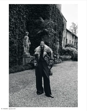 Model wearing a textured fur coat and wide-leg trousers standing outdoors on a gravel path near classical statues in a monochrome fashion editorial.