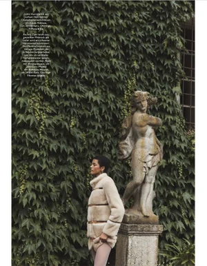 Model standing next to a stone classical statue in front of a wall covered in lush green ivy leaves.