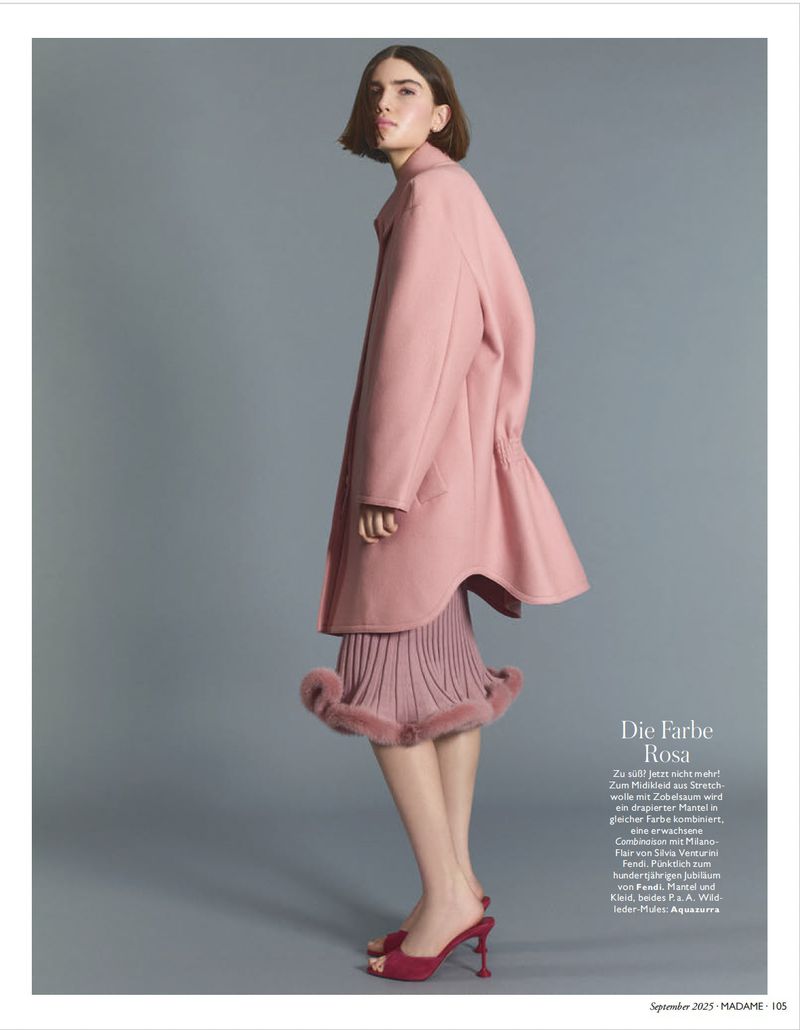 Elegant brunette woman poses in a blush-pink coat and matching skirt with fur trim against a gray background in a fashion editorial style.