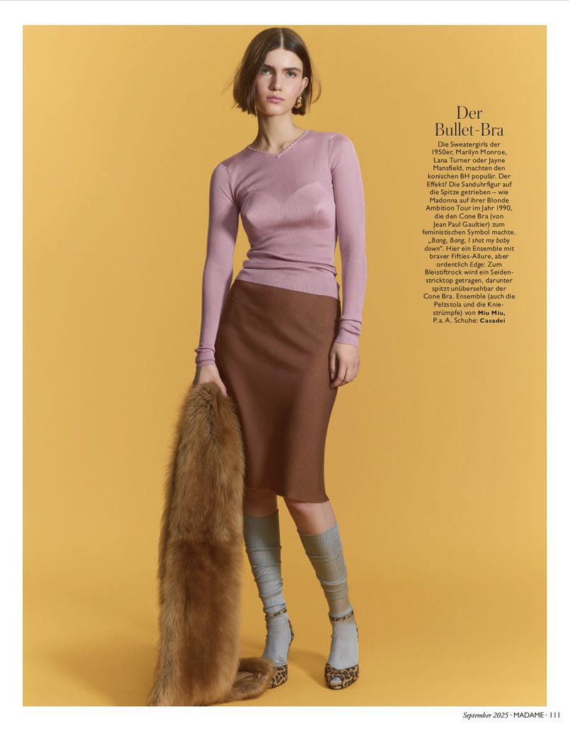 Full-length studio shot of a woman in pink top, brown skirt, knee-high socks, and heels, holding a fur stole against a yellow background.