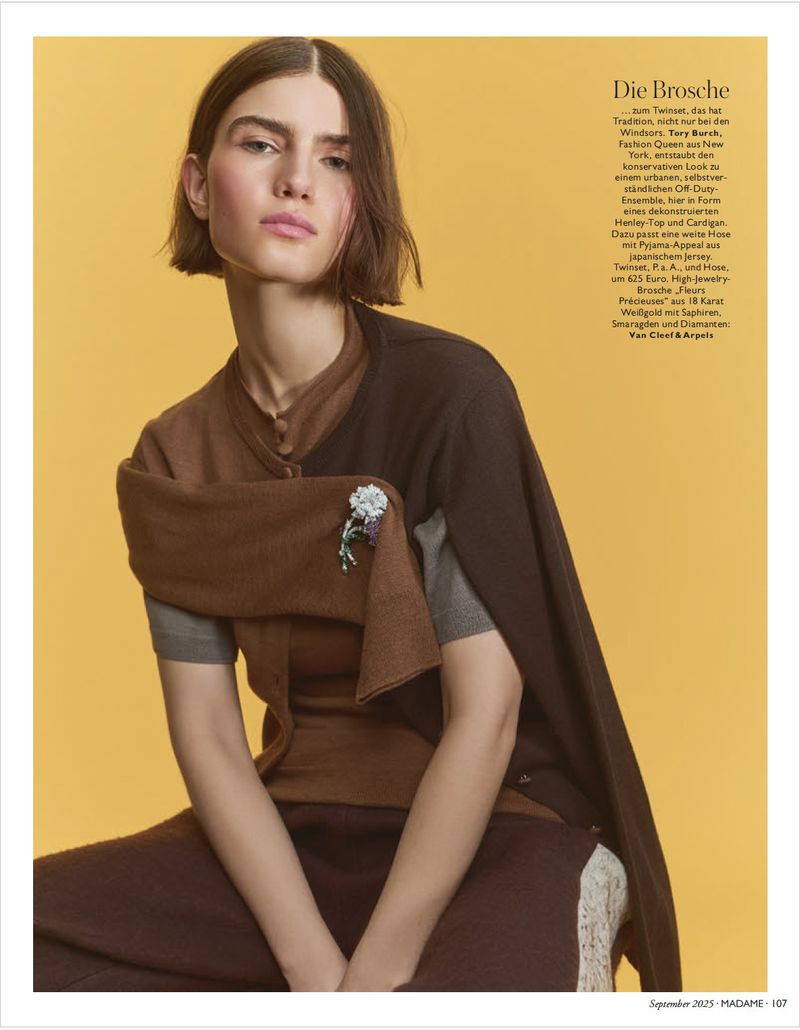 Elegant woman in brown and grey knitwear sitting against a yellow backdrop, showcasing a brooch in a fashion editorial shot.