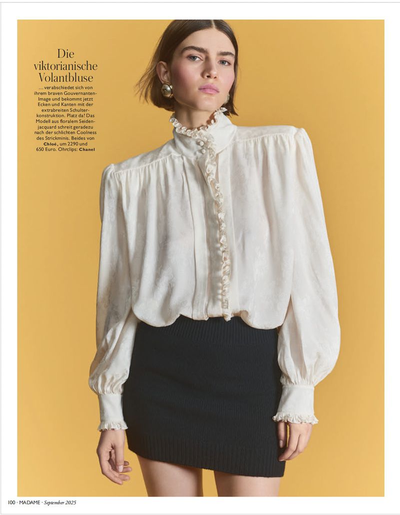 A woman in an off-white ruffled blouse and black miniskirt poses against a yellow backdrop in a fashion editorial shot.