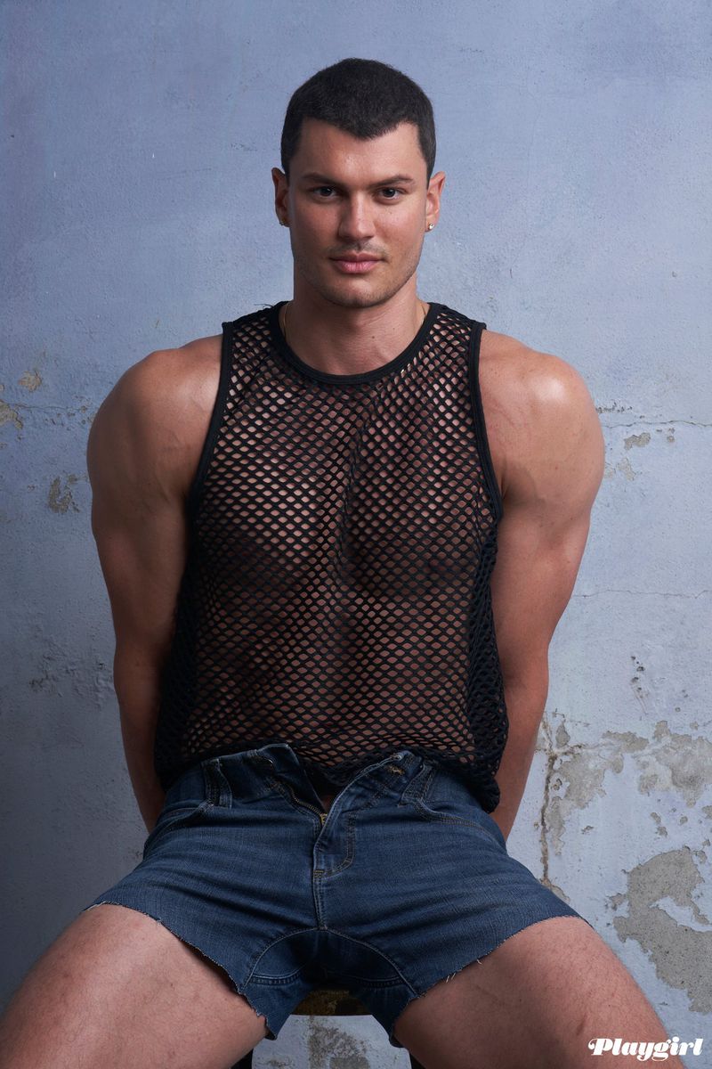 Portrait of a muscular adult male sitting on a stool wearing a black fishnet tank top and jean shorts in front of a textured wall.