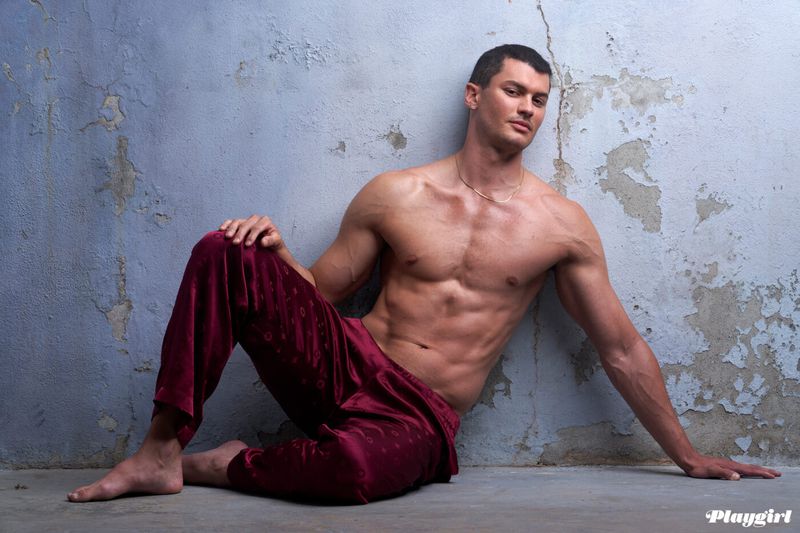 Shirtless man in patterned pants poses against cracked blue wall in studio lighting showcasing physique and fashion.