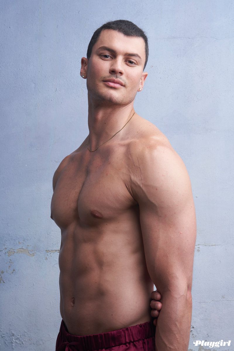Muscular shirtless man poses indoors against a textured gray wall. He wears maroon pants and faces forward with a neutral expression.
