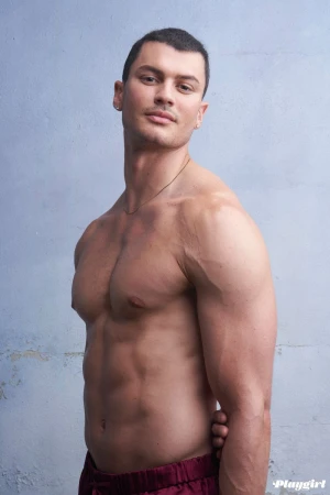 Muscular shirtless man poses indoors against a textured gray wall. He wears maroon pants and faces forward with a neutral expression.
