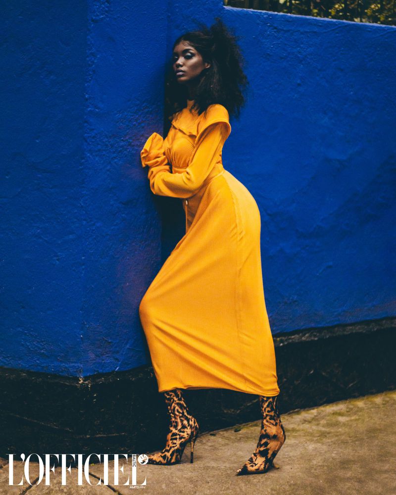 Dark-skinned woman in a yellow dress poses against a blue wall, wearing animal-print heels in an editorial fashion shot.