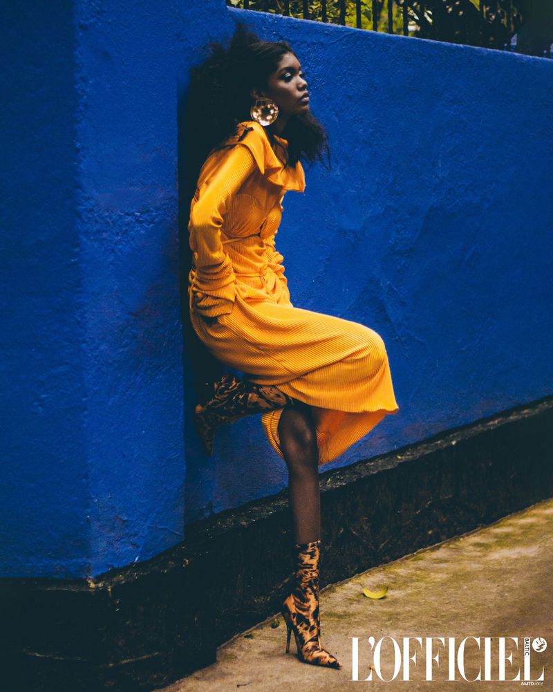 A stylish African woman poses in a yellow dress and snake-print boots against a blue wall, fashion editorial.