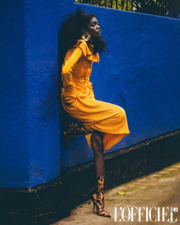 A stylish African woman poses in a yellow dress and snake-print boots against a blue wall, fashion editorial.