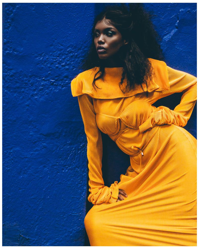 An African-American woman models a mustard-yellow dress against a textured blue wall, exuding fashion and style in a studio setting.