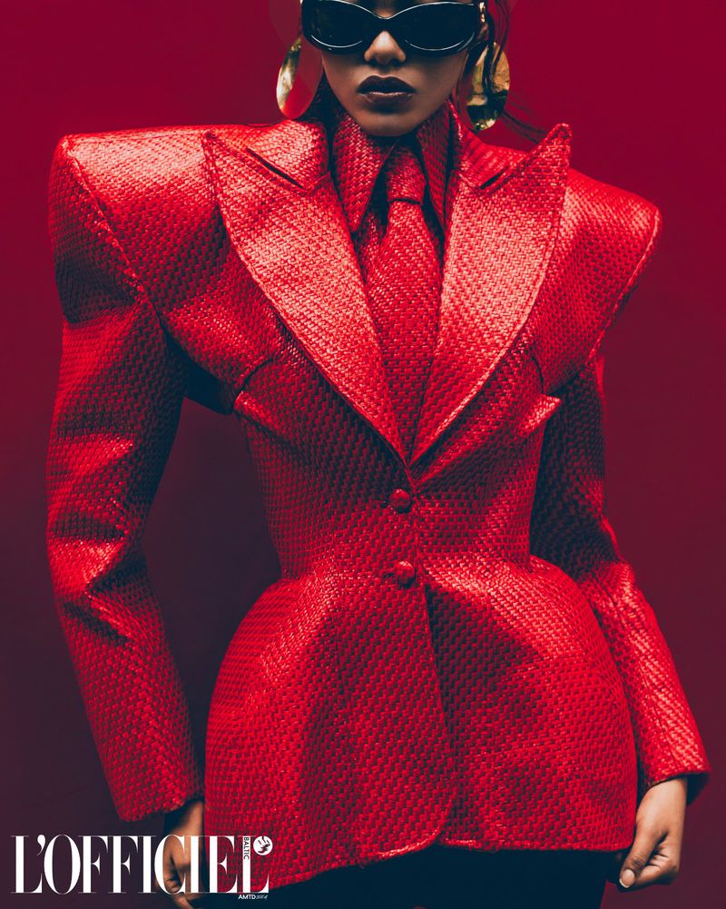 Editorial fashion shot of an adult female wearing a red blazer and tie with large gold earrings against a red gradient backdrop.