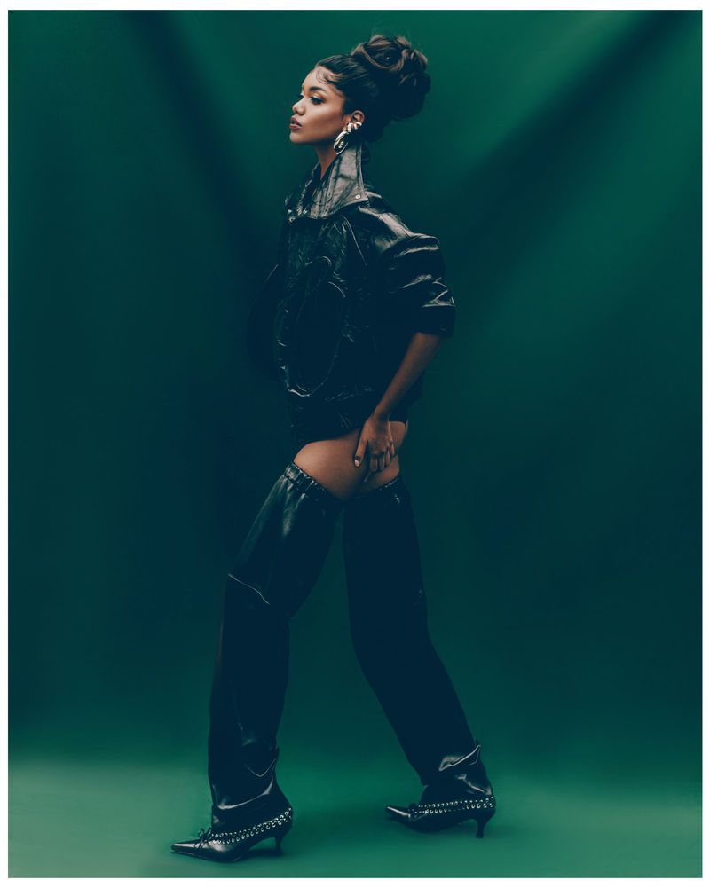 Profile shot of an adult female with styled hair wearing black leather-like clothing while posing against a dark green background.