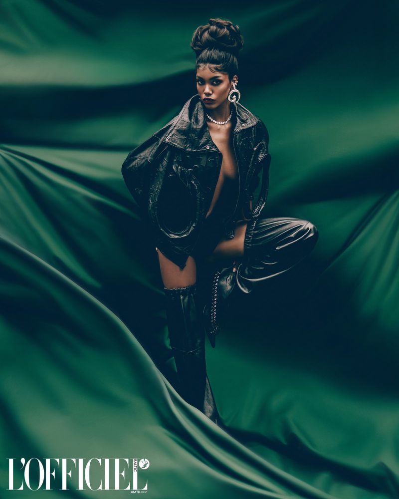 Fashion-forward female in a black jacket and thigh-high boots, posing against draped green fabric. Editorial style, magazine logo visible.