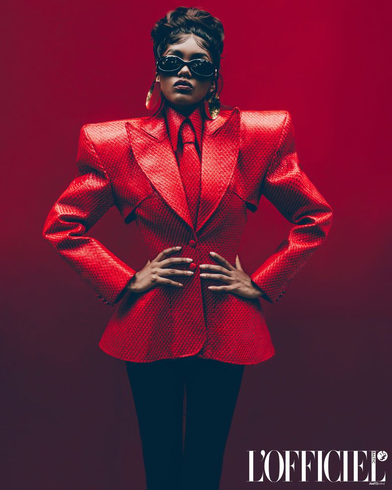 Portrait of a woman in a red suit with strong shoulder pads and sunglasses against a red gradient background, editorial fashion.