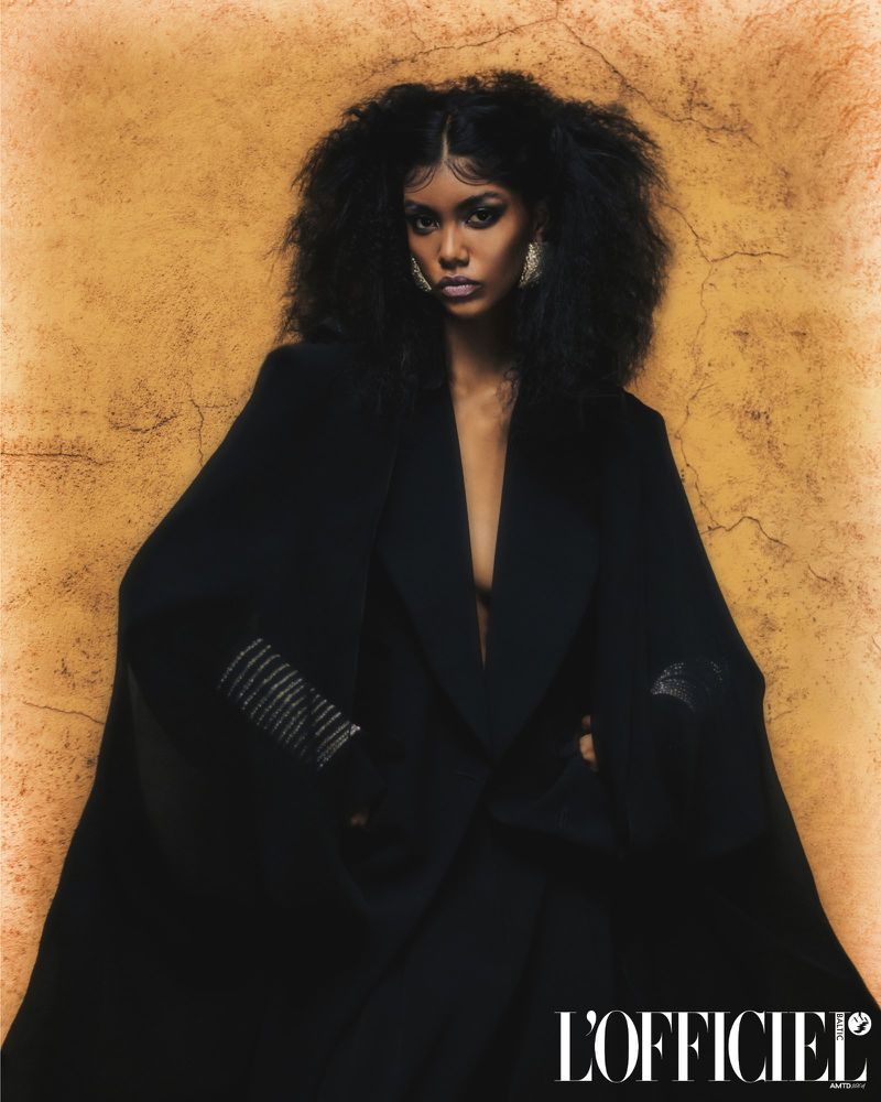 Portrait of a Black woman in a black coat against an ochre background. She has dark curly hair and is wearing large hoop earrings.