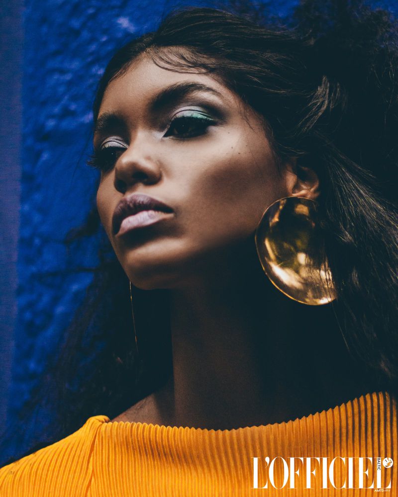 Close-up portrait of a Black female model with a yellow top against a textured blue background, featuring dramatic makeup and large earrings.