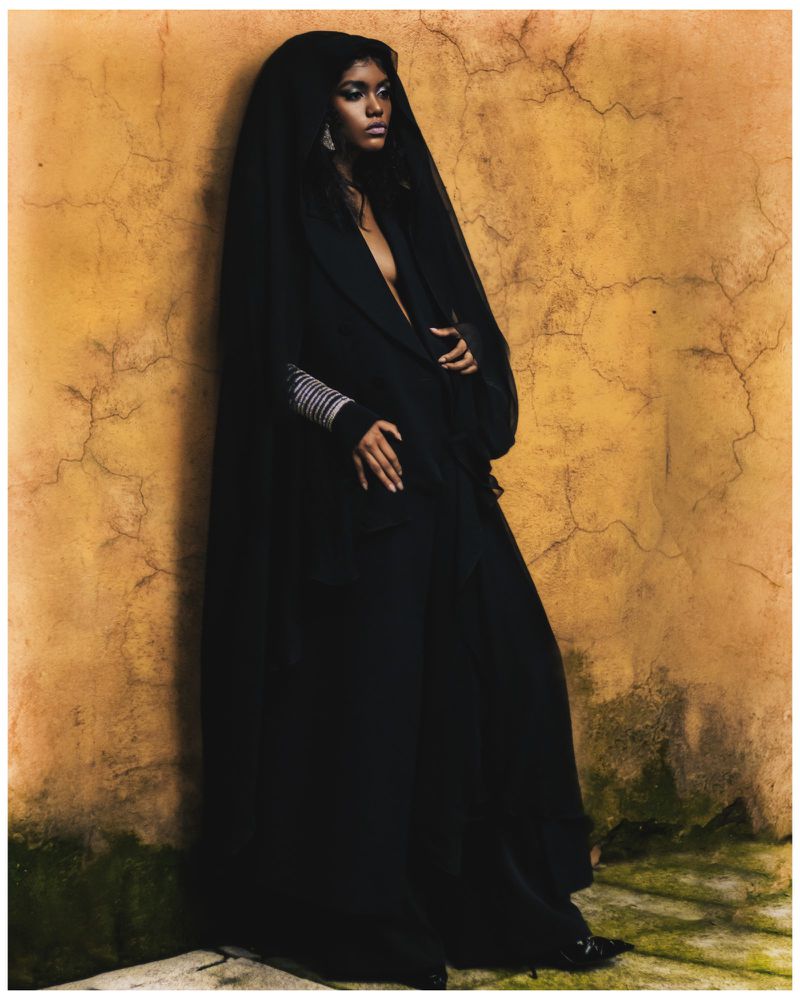 A Black woman stands in a black gown with a sheer hooded head covering against a tan cracked wall in a studio portrait.