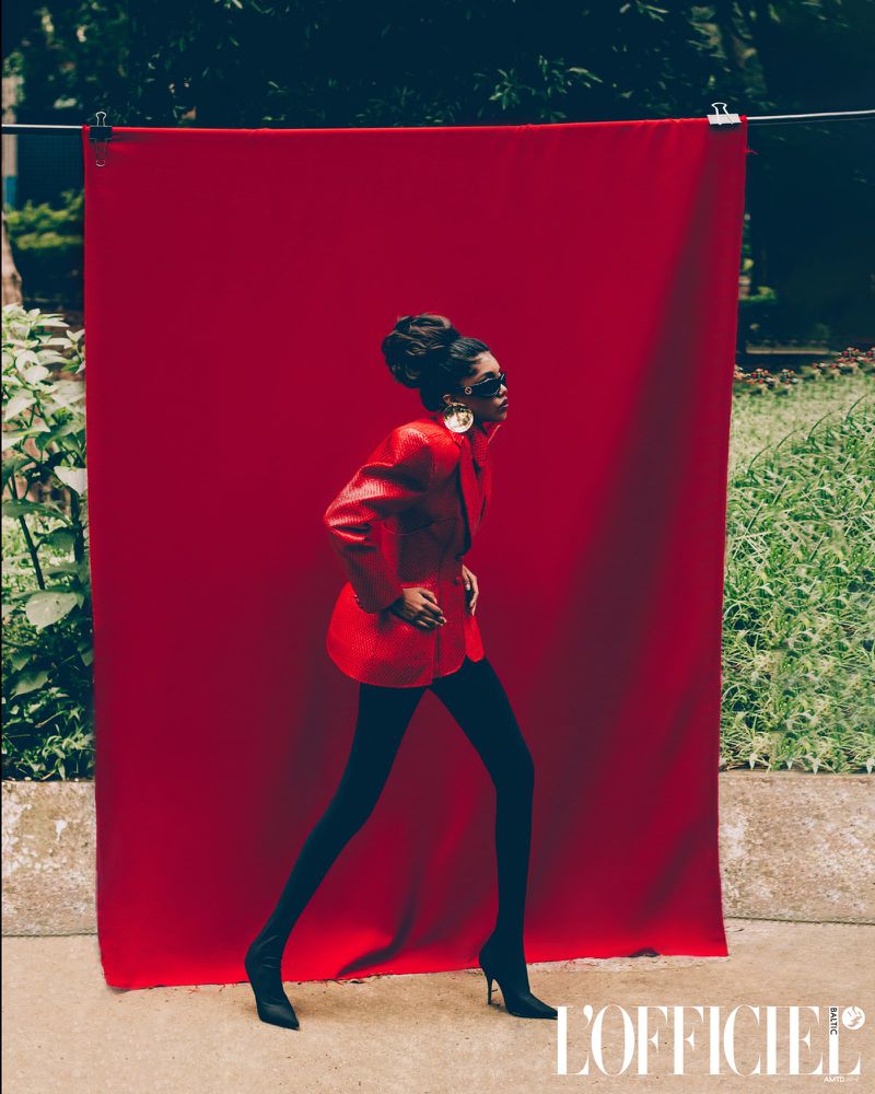 Fashionable woman in a red blazer and black tights walking against a red backdrop in an outdoor setting, style editorial.