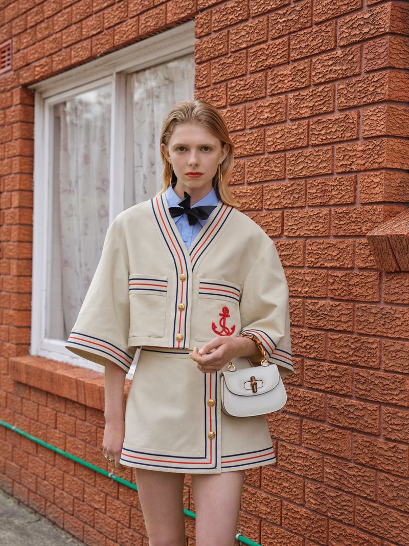 Young woman in a stylish cream-colored skirt suit with red and blue trim poses against a brick wall, holding a small white handbag.