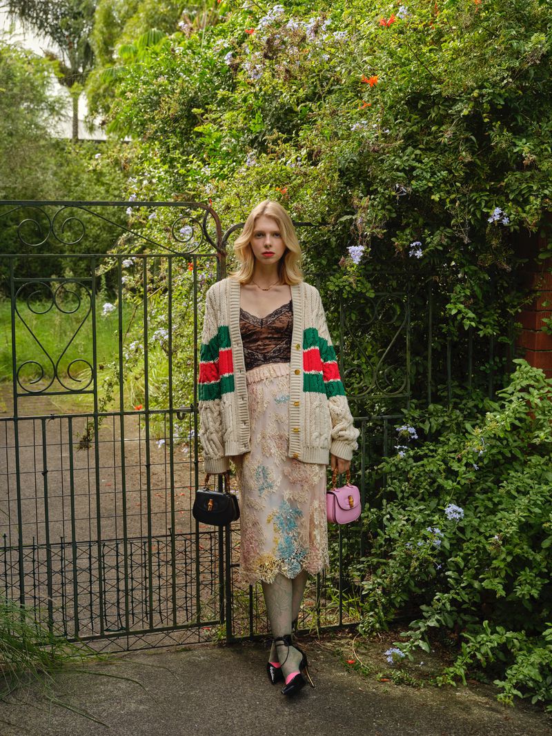 Model standing outdoors near a wrought iron gate wearing a striped cardigan, patterned skirt, and holding handbags.