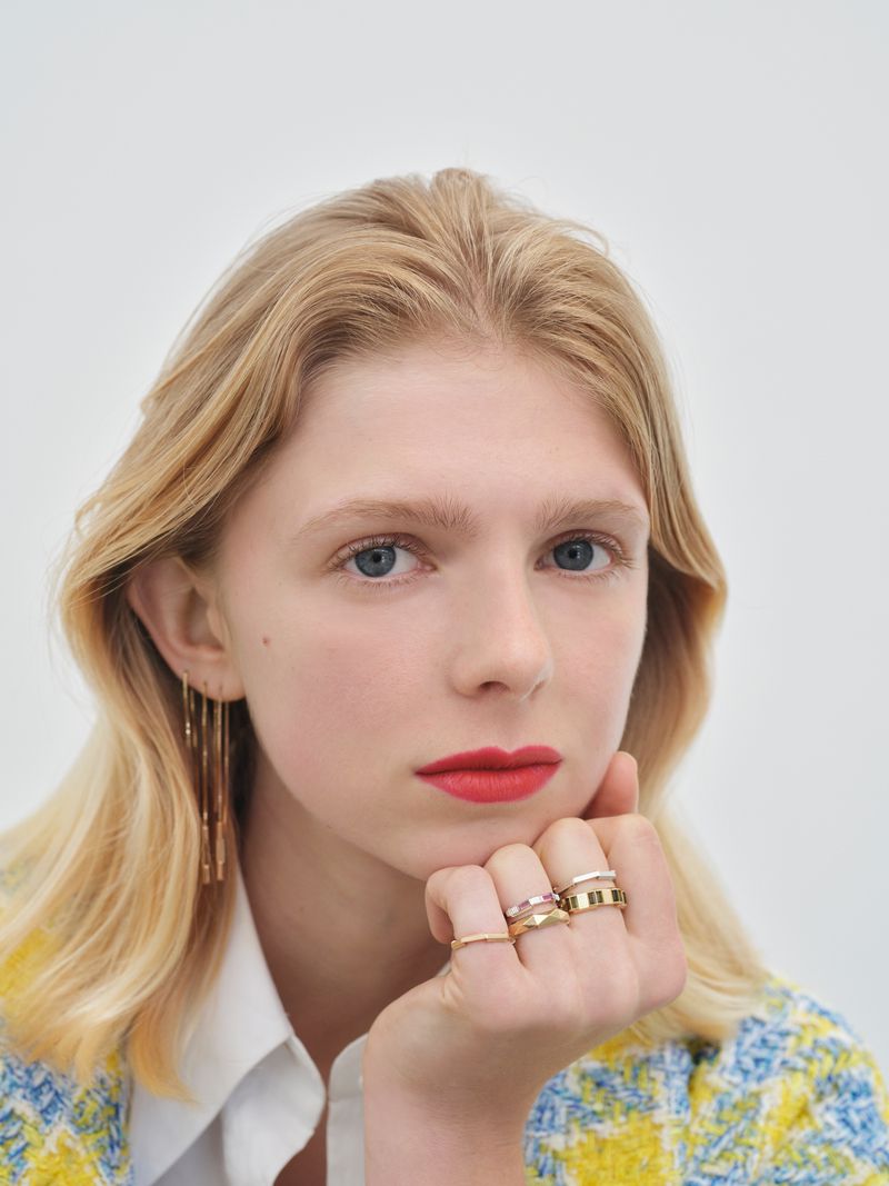 Close-up studio portrait of a blonde woman wearing gold rings, looking at the camera with red lipstick against a plain white background.
