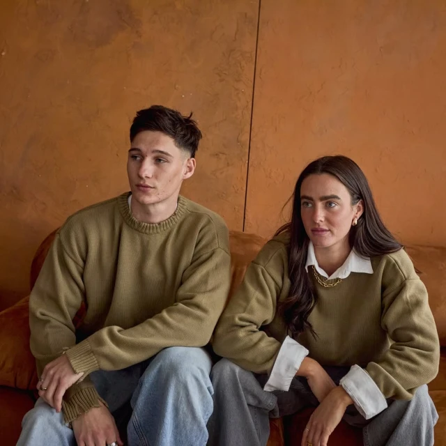 A young couple, a man and a woman, are sitting indoors wearing olive-green sweaters and jeans. They are posing with a neutral expression.