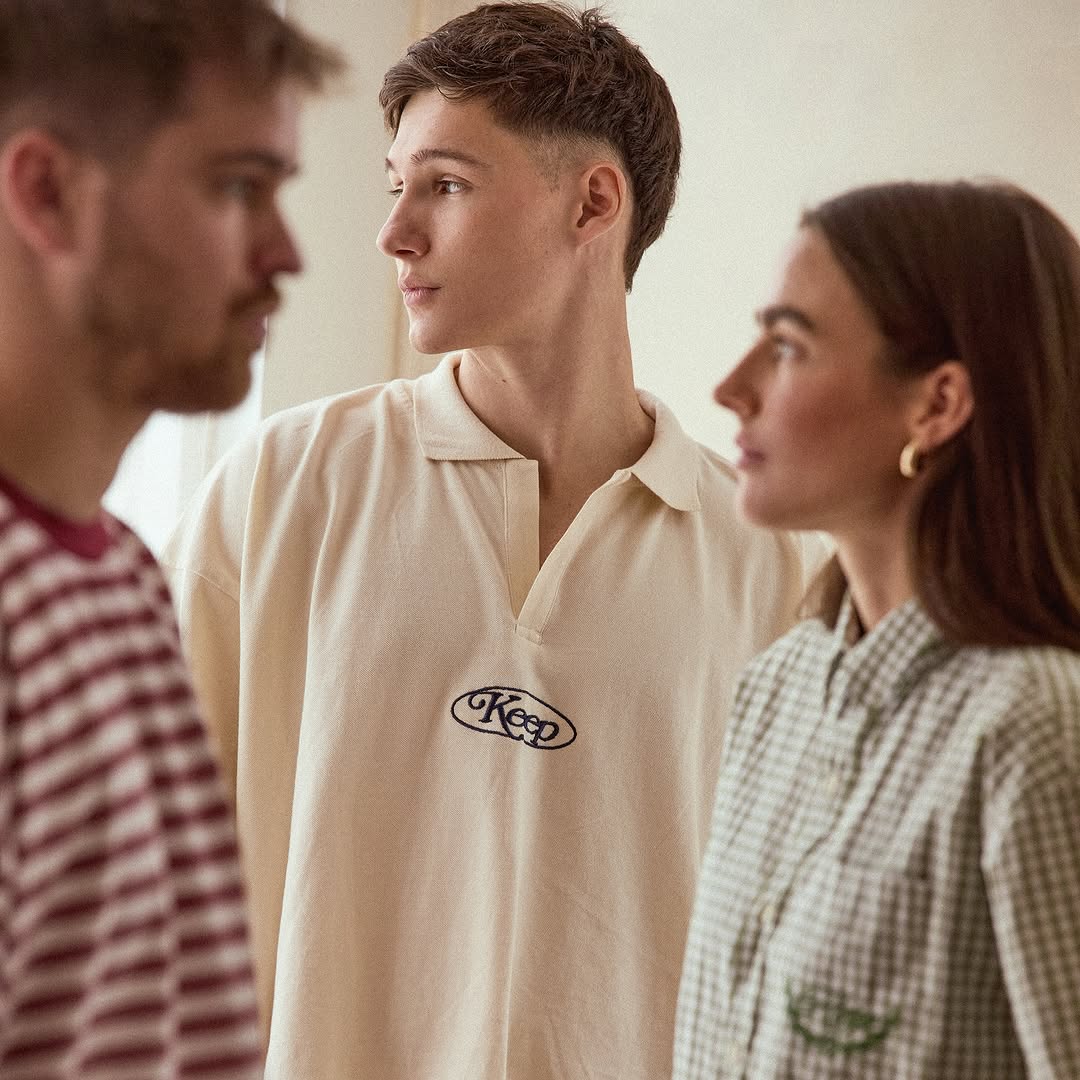 Three people stand indoors, wearing casual shirts in a medium studio shot. Two look toward the center figure in a cream top.