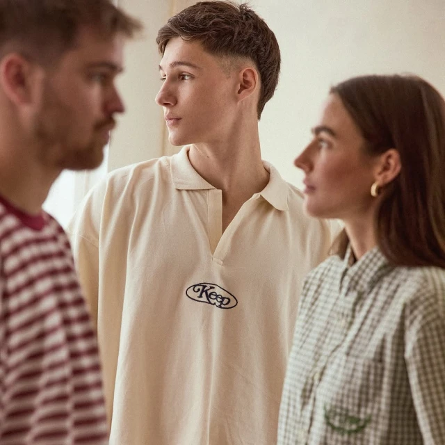 Three people stand indoors, wearing casual shirts in a medium studio shot. Two look toward the center figure in a cream top.