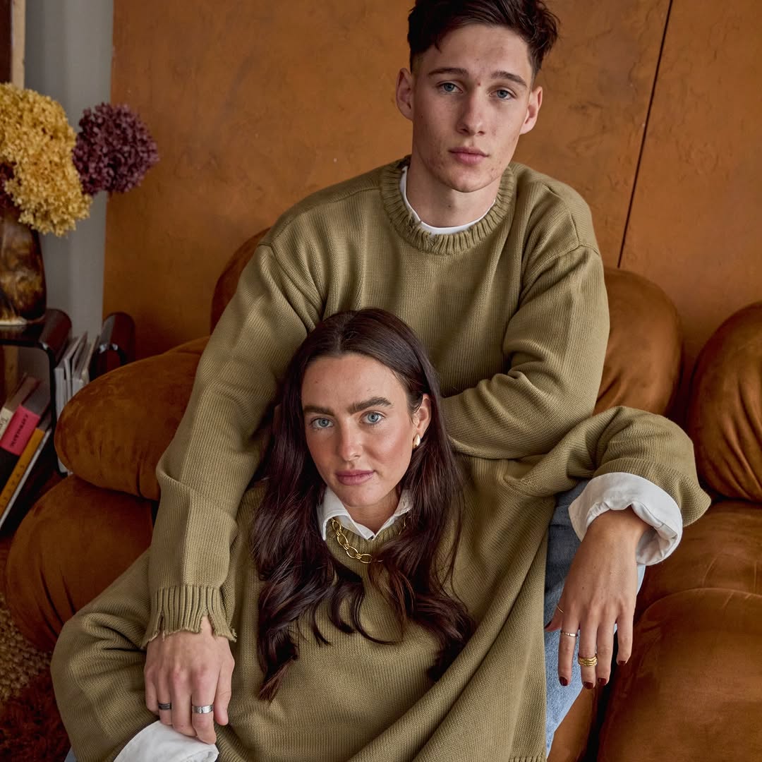 A young couple poses indoors on a brown sofa, both wearing olive green sweaters, with flowers and books in the background, looking at the camera.