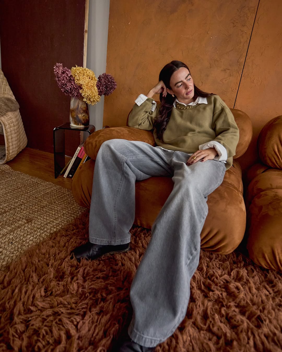 Stylish adult female sits in a brown chair in an indoor setting wearing wide leg jeans, and an olive green sweater with flowers on a table.