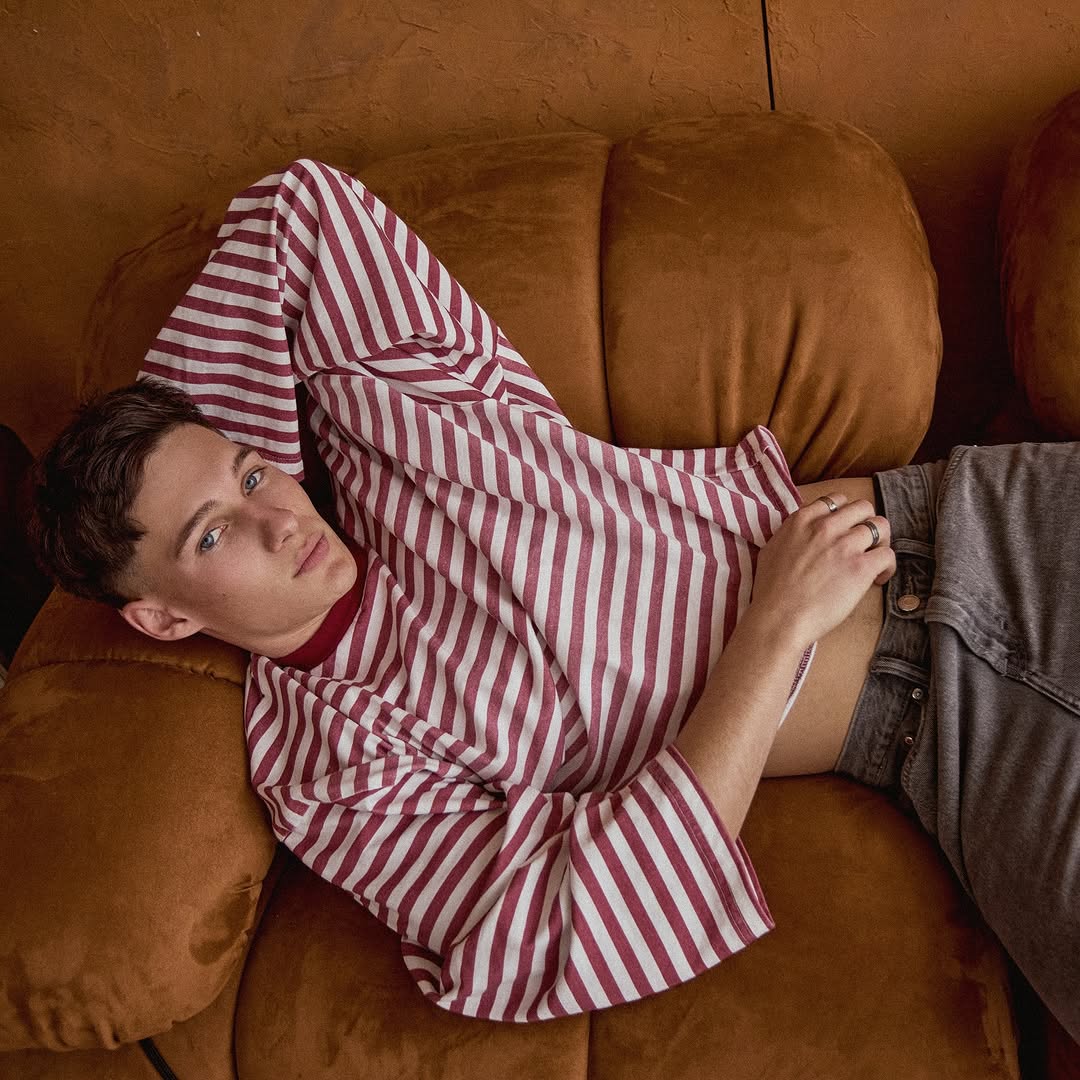 Young man relaxing on a brown couch. He is lying down wearing a striped shirt and jeans, looking at the camera indoors.