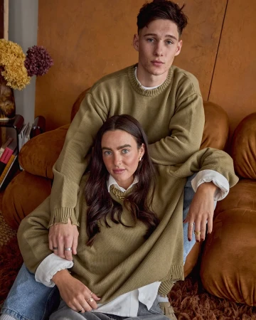 A young adult man and woman in a photo studio wear olive green sweaters; the man stands behind the seated woman in a medium shot.