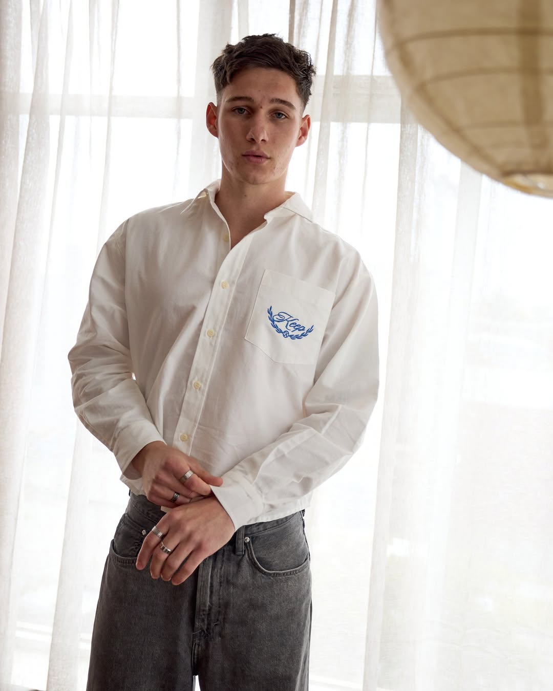 Portrait of a young adult man in a white button-down shirt and grey jeans, standing indoors with soft window light and a relaxed pose.