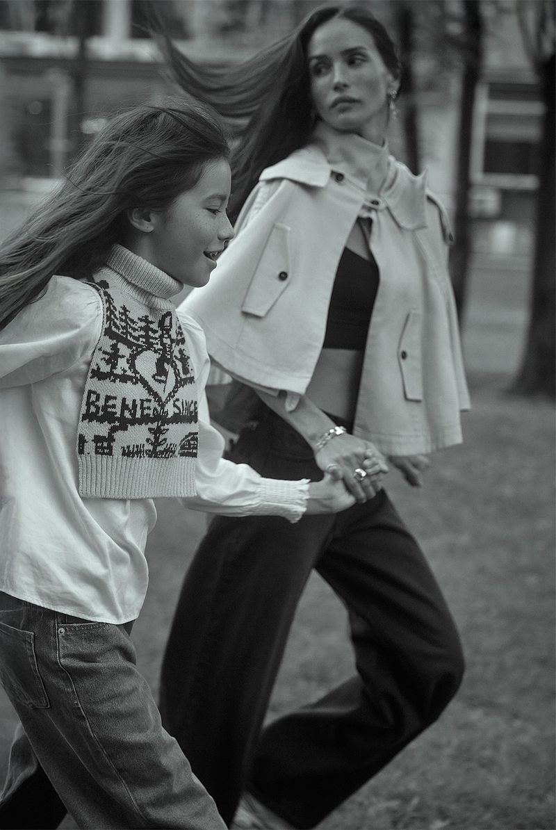 Black and white shot of a mother and daughter holding hands running in a park. The woman wears a cape and jeans; the girl wears jeans and a sweater vest.