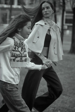 Black and white shot of a mother and daughter holding hands running in a park. The woman wears a cape and jeans; the girl wears jeans and a sweater vest.