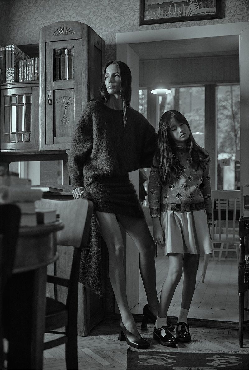 Monochrome portrait of an adult woman and a child standing indoors. The woman wears a dark sweater dress, while the child wears a dress with a pattern.