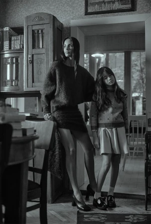 Monochrome portrait of an adult woman and a child standing indoors. The woman wears a dark sweater dress, while the child wears a dress with a pattern.