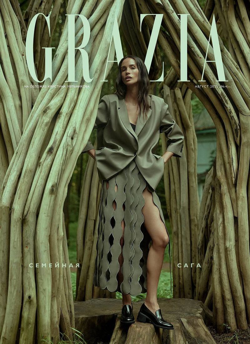Fashion editorial cover featuring a woman in an olive-green suit and skirt posing in front of a wooden structure in a forest setting.