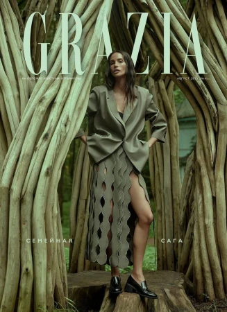 Fashion editorial cover featuring a woman in an olive-green suit and skirt posing in front of a wooden structure in a forest setting.