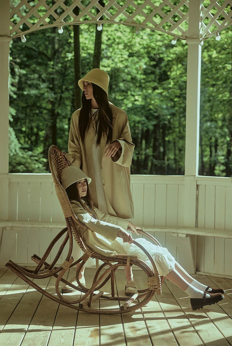 Two women pose in a gazebo. One is seated in a wicker rocking chair, while the other stands nearby. Both wear matching bucket hats and coats.
